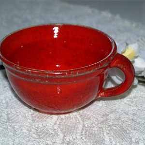 Könnte beinhalten: Eine rote Keramik-Teetasse mit leicht strukturierter Oberfläche. Die Tasse hat eine abgerundete Form und einen Henkel.