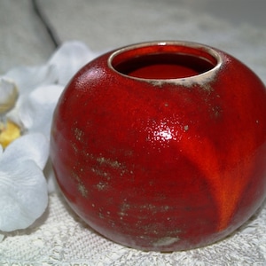 Op de afbeelding: Een kleine, ronde, rode keramische vaas met een glanzende afwerking. De vaas heeft een brede opening en een licht gestructureerd oppervlak. Op de achtergrond is een witte orchidee bloem te zien. De vaas wordt waarschijnlijk gebruikt voor decoratieve doeleinden.