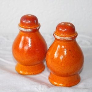 Op de afbeelding: Een paar oranje keramische zout- en peperstrooiers met een glanzende afwerking en witte randen. De strooiers hebben een bolle vorm en kleine, ronde deksels. Ze staan op een wit oppervlak. Hoogte ongeveer 8 cm.