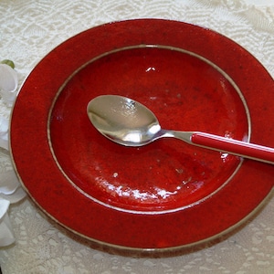 Może przedstawiać: Czerwony ceramiczny talerz z srebrną obwódką i srebrną łyżką na górze. Talerz jest pusty.