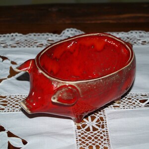 May include: A red ceramic piggy bank with a glazed finish. The piggy bank is shaped like a pig with a rounded belly and four legs.