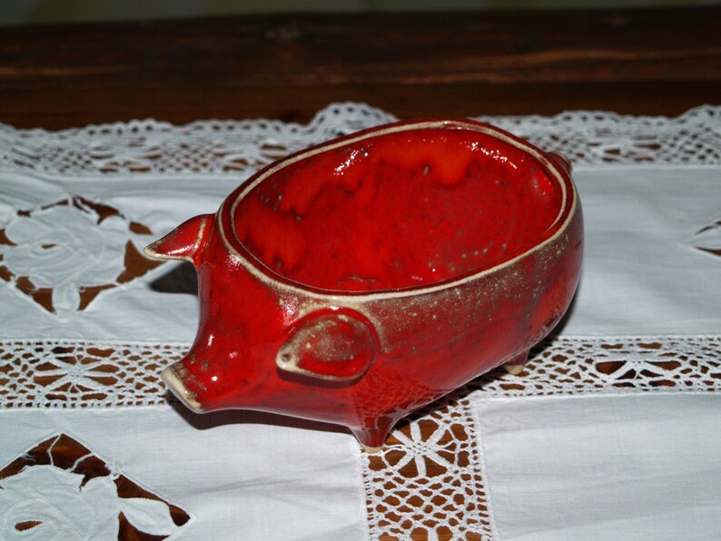 May include: A red ceramic piggy bank with a glazed finish. The piggy bank is shaped like a pig with a rounded belly and four legs.
