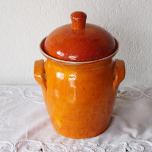 Può includere: Un vaso di ceramica smaltata con coperchio. Il vaso è di un vibrante colore arancione, con un coperchio e un manico marrone rossiccio più scuro. Il vaso ha due manici laterali e un piccolo pomello sul coperchio. Il vaso è appoggiato su una superficie bianca.