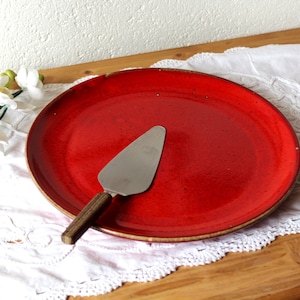 Può includere: Un piatto da portata in ceramica rosso vivo con una paletta per torte abbinata. Il piatto ha una superficie leggermente macchiata e un bordo marrone dorato. La paletta per torte ha una lama in acciaio inossidabile e un manico in legno. Su una tovaglia di pizzo bianco.