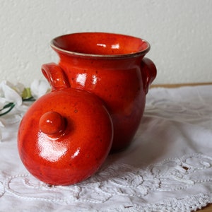 Op de afbeelding: Een glanzende, oranje keramische pot met een bijpassende deksel. De pot heeft twee kleine handvatten en een licht uitlopende rand. De deksel is rond met een kleine knop. De pot en deksel staan op een witte, geborduurde doek.