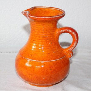 Könnte beinhalten: Orangefarbener Keramik Krug mit breiter Öffnung und Henkel. Der Krug hat eine strukturierte Oberfläche und eine glänzende Oberfläche.