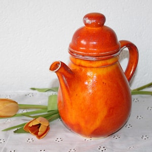 Könnte beinhalten: Orangefarbene Teekanne aus Keramik mit Deckel und Henkel. Die Teekanne steht auf einer weißen Tischdecke mit Blumenmuster.