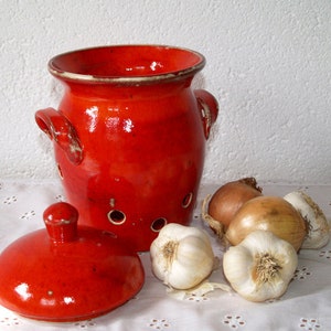 Pode incluir: Um recipiente de cerâmica vermelha para guardar cebolas e alho, com tampa. O recipiente tem furos para ventilação e está sobre uma toalha de mesa branca com cebolas e alho ao redor.