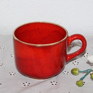Könnte beinhalten: Eine rote Keramiktasse mit weißem Rand. Die Tasse steht auf einer weißen Tischdecke mit Blumenmuster.