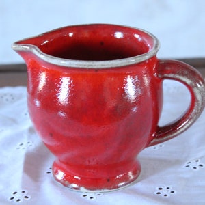 Può includere: Un'anfora in ceramica rossa con un bordo bianco e un manico bianco. L'anfora ha una finitura lucida ed è appoggiata su un tovagliolo di pizzo bianco.