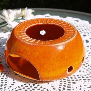 Può includere: Portacandele per tealight in ceramica arancione con forma rotonda e un foro al centro. Il portacandele ha una superficie testurizzata e un piccolo foro sul lato.