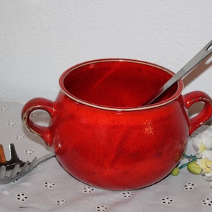 Könnte beinhalten: Eine rote Suppenterrine aus Keramik mit zwei Henkeln und einer silbernen Schöpfkelle im Inneren. Die Terrine steht auf einer weißen Spitzentischdecke.