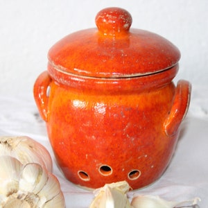 Puede incluir: Un tarro de cerámica para ajos con tapa, acabado en un vibrante esmalte naranja. El tarro tiene forma redonda con dos asas y tres orificios de ventilación. Se ven bulbos de ajo en primer plano. Este artículo de cocina está diseñado para guardar ajos.