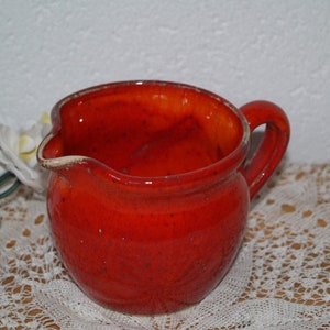 Può includere: Un'anfora in ceramica rossa con corpo arrotondato e manico. L'anfora ha una smaltatura macchiata e una superficie leggermente irregolare.