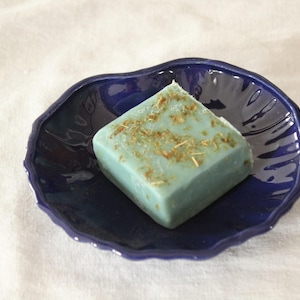 Può includere: Una saponetta quadrata, azzurra, con erbe secche in cima, appoggiata su un piatto di ceramica blu scuro a forma di foglia. La saponetta e il piatto sono su sfondo bianco.