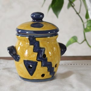 Puede incluir: Un tarro de cerámica amarillo con tapa, adornado con patrones y acentos geométricos azul marino. El tarro tiene pequeñas asas y agujeros decorativos. Esta pieza artesanal es adecuada para la cocina o la decoración del hogar.
