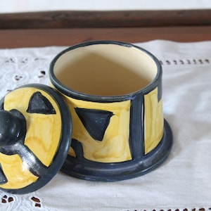 Può includere: Un vaso di ceramica con coperchio, dipinto con motivi geometrici gialli, blu e neri. Il vaso ha un pomo sul coperchio.