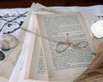 May include: A bundle of old book pages tied with string, showing printed text. The pages are arranged with a seashell, a small stone, and white roses on a lace-like cloth.