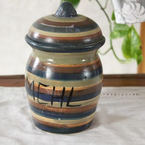 Op de afbeelding: Een gestreepte keramische pot met deksel, in bruin, blauw en groen. Het woord "MEHL" staat in het zwart geschreven. De pot is waarschijnlijk bedoeld voor het bewaren van droge waren.