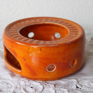 Peut inclure: Un chauffe-thé en céramique orange de forme ronde et à la finition brillante. Le chauffe-thé présente un design circulaire avec une série de trous et un bord texturé. Il mesure environ 10 cm de diamètre.