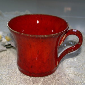 Könnte beinhalten: Eine leuchtend rote Keramiktasse mit gebogenem Henkel. Die Tasse hat eine glänzende Oberfläche und eine leicht strukturierte Oberfläche. Der Rand und die Kanten des Griffs haben eine kontrastierende braune Umrandung. Ein dekorativer Artikel für die Küche.