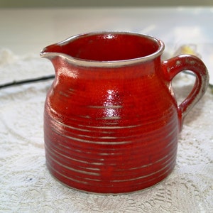 Può includere: Brocca in ceramica rossa fatta a mano, con manico e beccuccio. La brocca ha una superficie strutturata con linee orizzontali e un bordo chiaro. L'interno è anch'esso rosso.