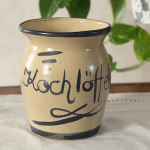 Puede incluir: Un recipiente de cerámica beige para utensilios de cocina con borde y base azul oscuro. Las palabras "Kochlöffel" están escritas en escritura azul oscuro en la parte delantera. El recipiente tiene una forma ligeramente acampanada.