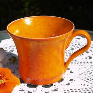 Puede incluir: Una taza de cerámica naranja hecha a mano con un asa curvada. La taza tiene una textura moteada y un acabado brillante. La taza está sobre un mantel individual de encaje blanco, con una flor naranja y flores blancas en el fondo.