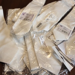 Könnte beinhalten: Eine Ansammlung weißer Krawatten, einige noch in ihrer Originalverpackung, liegt auf einer dunklen Holzoberfläche. Die Krawatten scheinen aus einem glatten Material zu sein. Einige haben Etiketten.