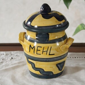 Op de afbeelding: Een gele en marineblauwe keramische pot met deksel. De cilindervormige pot heeft handvatten en het woord 'MEHL' in zwarte letters. Het deksel is bekroond met een kleine knop.