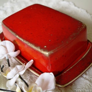 Op de afbeelding: Een rechthoekige rode keramische botervloot met bijpassend deksel. De schaal heeft een gouden rand langs de randen. De botervloot staat op een wit kanten oppervlak, met witte bloemen op de voorgrond.