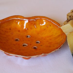 Könnte beinhalten: Orangefarbene Keramik-Seifenschale mit Abflusslöchern, neben mehreren handgemachten Seifenstücken. Die Schale hat eine glänzende Oberfläche und eine einzigartige Form. Die Seife scheint natürliche Inhaltsstoffe zu haben.