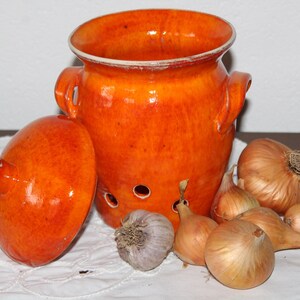 Könnte beinhalten: Ein orangefarbener Keramik-Zwiebelbehälter mit Deckel und Belüftungslöchern. Der Behälter steht neben einer Sammlung von Zwiebeln und einer Knoblauchzehe, alles auf einer weißen Spitzentischdecke.