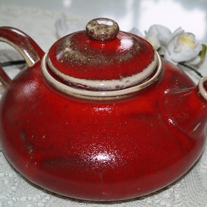 Può includere: Una teiera in ceramica rossa con un bordo bianco e un coperchio. La teiera ha una glassa macchiata e un aspetto rustico.