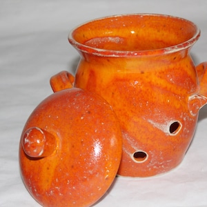 Könnte beinhalten: Orangefarbene Keramikdose mit Deckel und Löchern an der Seite. Die Dose hat eine gesprenkelte Glasur.