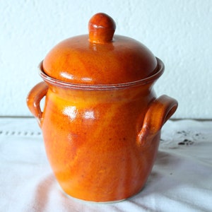 Könnte beinhalten: Ein Keramiktopf mit Deckel und glänzender orangefarbener Glasur. Der Topf hat zwei Griffe und eine runde Form, ideal für die Küche. Der Deckel hat einen kleinen Knauf. Der Topf steht vor einem weißen Hintergrund.