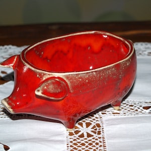 May include: A red ceramic pig-shaped bowl with a glazed finish. The bowl has a rounded shape and four short legs.