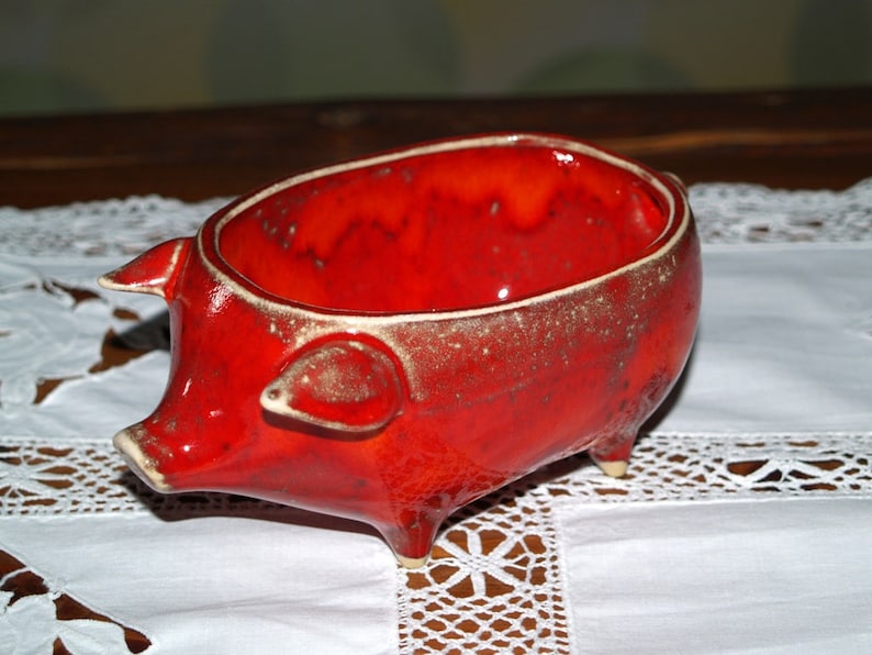 May include: A red ceramic pig-shaped bowl with a glazed finish. The bowl has a rounded shape and four short legs.