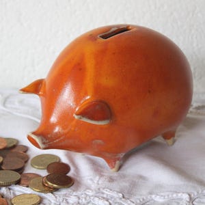 Könnte beinhalten: Ein Sparschwein aus Keramik in Orange- und Gelbtönen mit einem Münzschlitz oben. Das Schwein hat kleine Ohren, eine Schnauze und winzige Beine. Daneben liegt ein Haufen Münzen, was auf Ersparnisse hindeutet. Das Sparschwein ist ein klassischer Artikel zum Sparen.