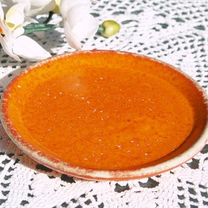 Könnte beinhalten: Eine kleine, runde Keramikschale mit leuchtend orangefarbener Glasur. Die Schale hat einen leicht erhöhten Rand mit einem rotbraunen Rand. Das Innere der Schale hat ein gesprenkeltes Aussehen. Die Schale steht auf einer weißen, spitzenartigen Oberfläche.