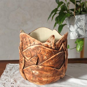 Può includere: Fioriera in ceramica fatta a mano con un design unico di pesci in rilievo. La fioriera è di colore marrone caldo con un interno color crema. I pesci sono finemente dettagliati, aggiungendo un tocco rustico e artistico all'arredamento.