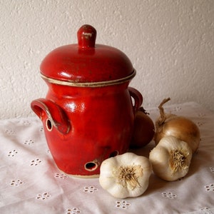 Op de afbeelding: Een rode keramische knoflookpot met een deksel en ventilatiegaten. De pot staat op een wit tafelkleed met drie teentjes knoflook en een ui.