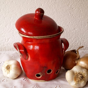 May include: Red ceramic garlic keeper with a lid and holes for ventilation. The keeper is surrounded by garlic bulbs and onions.