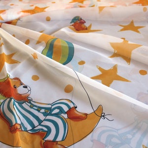 Può includere: Tenda bianca trasparente con un motivo giallo, arancione e blu di stelle, punti e un orsacchiotto che tiene un palloncino. L'orsacchiotto indossa un pigiama a righe blu e bianche ed è seduto su una luna crescente gialla.