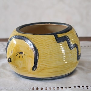 Könnte beinhalten: Ein gelber Keramiktopf mit einem schwarz-blauen bemalten Design eines Hundefaces. Der Topf hat eine abgerundete Form und eine breite Öffnung.