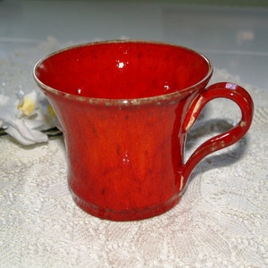 Peut inclure: Une tasse en céramique rouge vif avec une finition brillante et marbrée, dotée d'un bord large et évasé. La tasse présente une anse incurvée sur le côté droit, avec des variations subtiles dans le vernis rouge et une surface légèrement texturée.