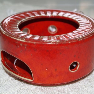 May include: Red ceramic tea warmer with a round opening and a handle on the side. The warmer has several small holes around the base.