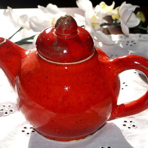 Pode incluir: Uma pequena chaleira de cerâmica vermelha com corpo arredondado e tampa. A chaleira tem uma alça e um bico.