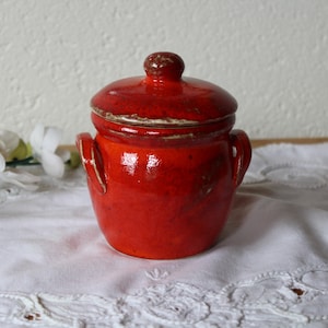 Op de afbeelding: Een kleine, rode keramische pot met een deksel. De pot heeft twee handvatten aan de zijkanten en een licht afgeronde vorm. Het deksel is licht gewelfd en heeft een kleine knop bovenop.