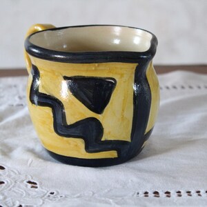 Può includere: Una tazza in ceramica gialla con disegni geometrici neri. La tazza ha una forma arrotondata e un manico.